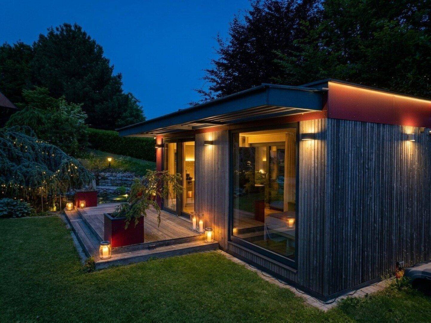 Modernes Nebengebäude mit Holzterrasse, Rasenfläche und stimmungsvoller Beleuchtung im Garten.