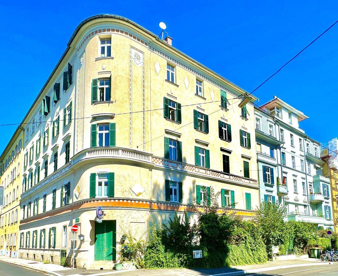 Historisches Eckgebäude mit gelber Fassade, grünen Fensterläden und dekorativen Elementen unter blauem Himmel.