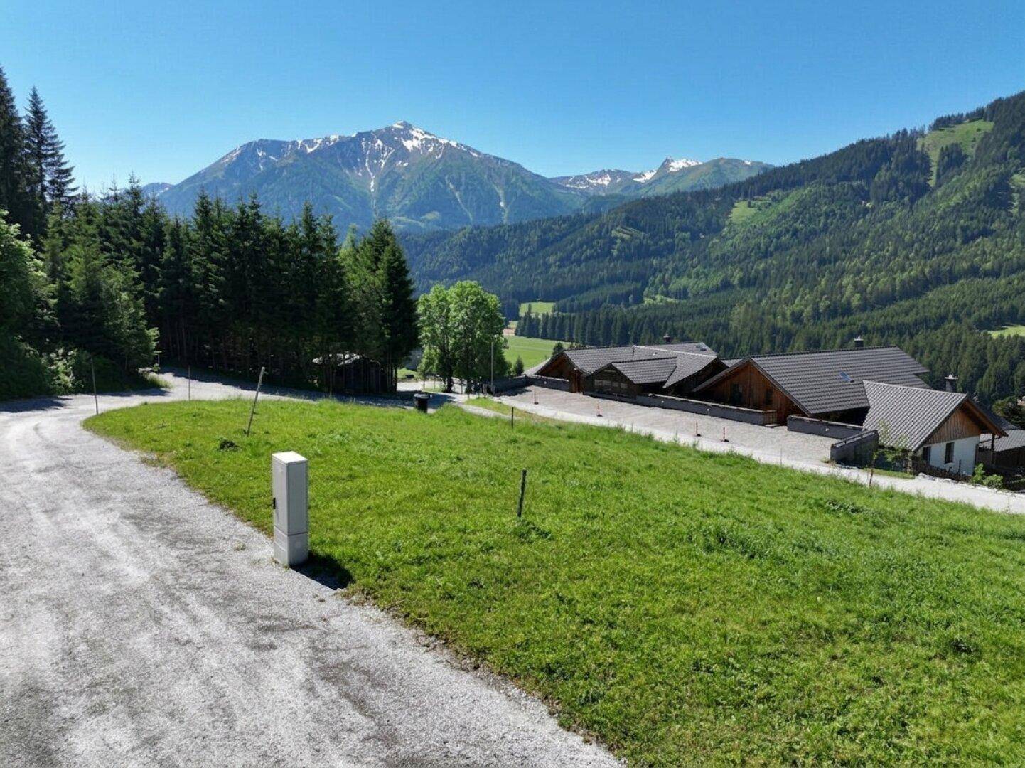 Malerische Berglandschaft mit grünen Wiesen, Schotterweg und traditionellen Häusern unter blauem Himmel.