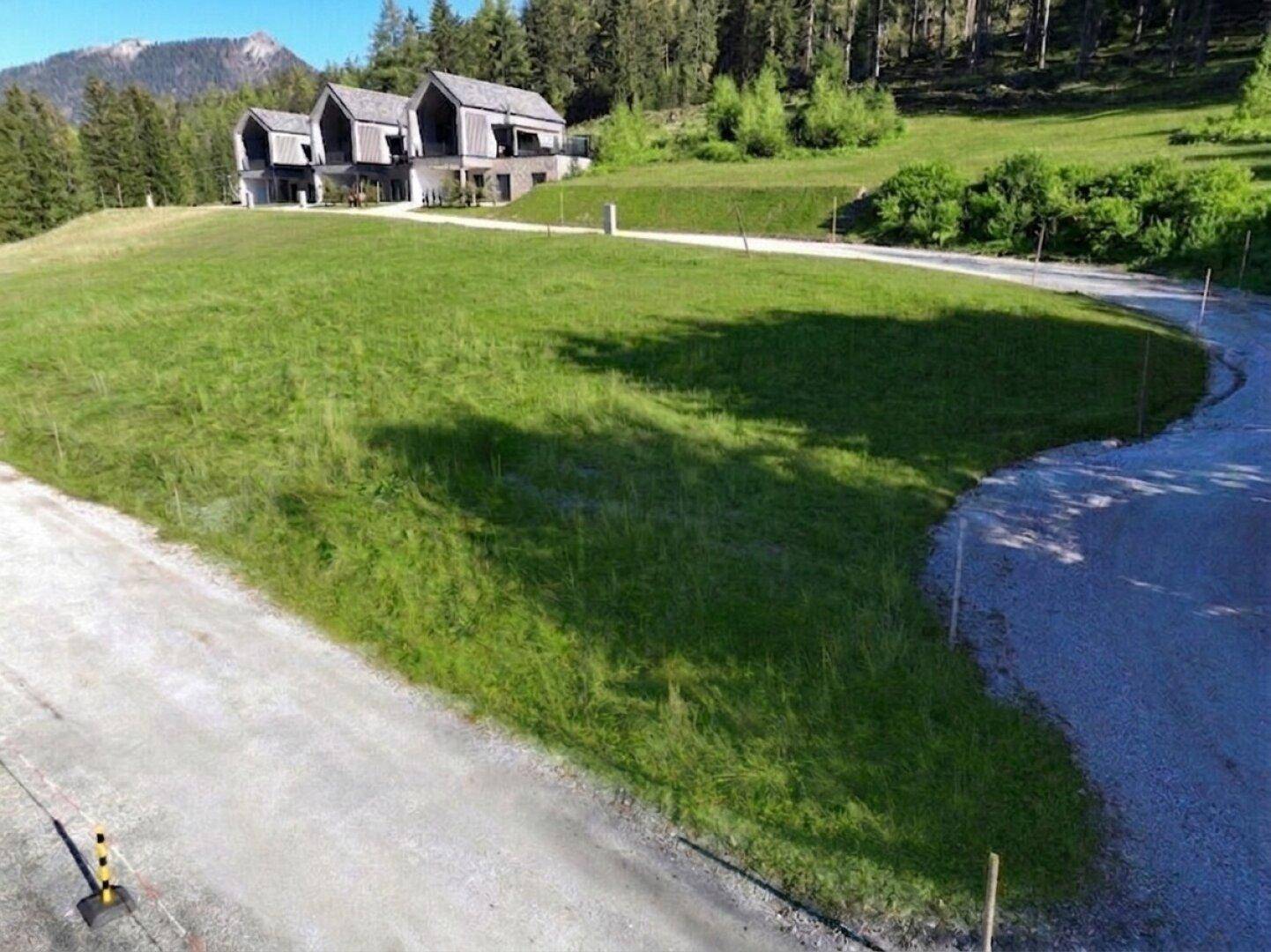Grüne Wiese mit Schotterweg und modernen Häusern in der Ferne, umgeben von Bergen und Wald.
