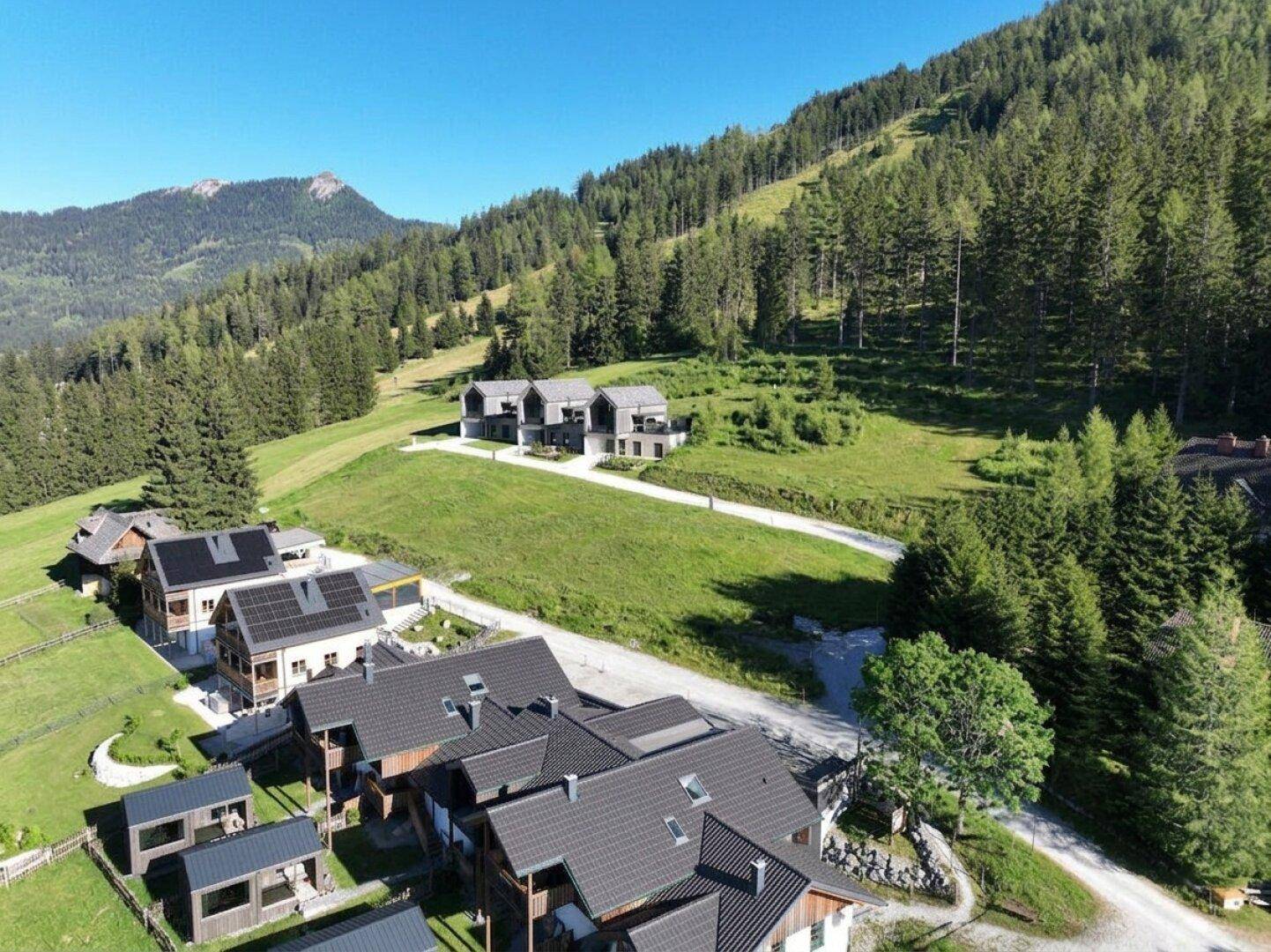 Luftaufnahme einer Siedlung moderner Häuser in einem grünen Tal, umgeben von bewaldeten Bergen.