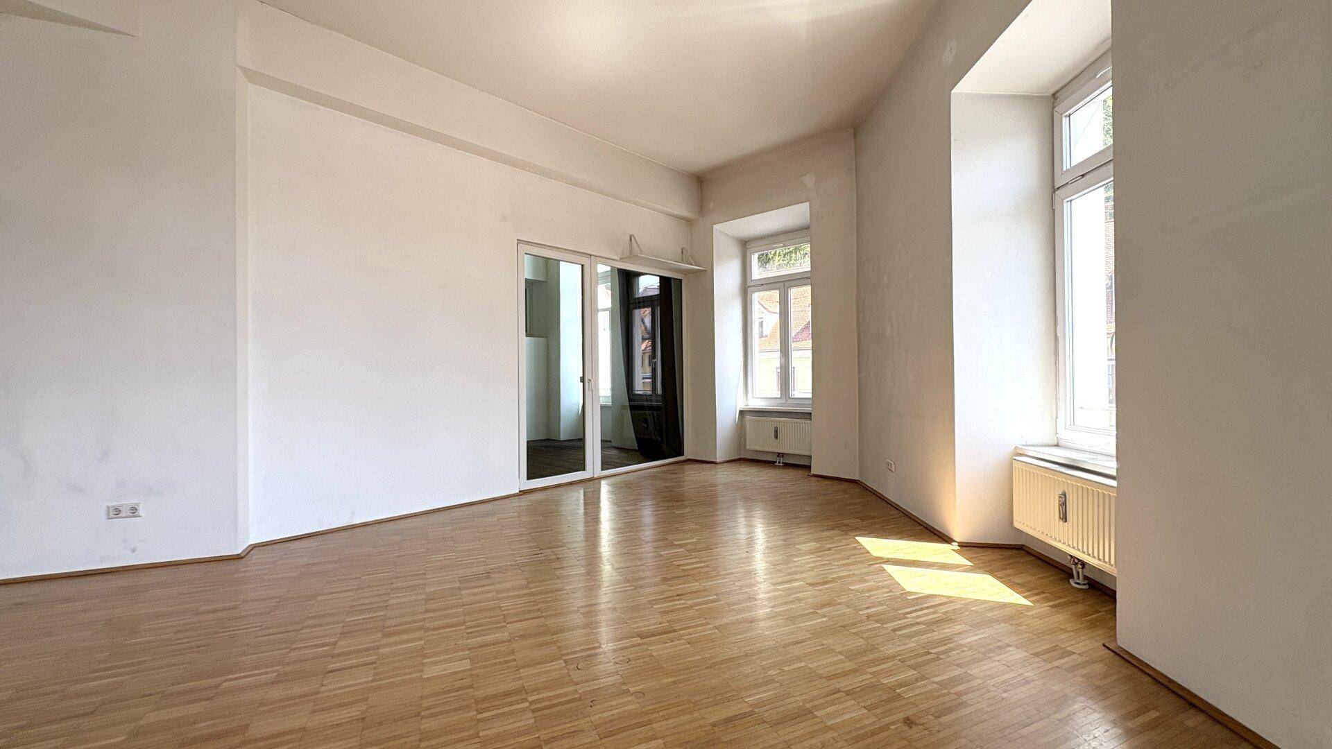 Geräumiges Wohnzimmer mit Parkettboden und einer Glasschiebetür, die zu einem weiteren Raum führt.