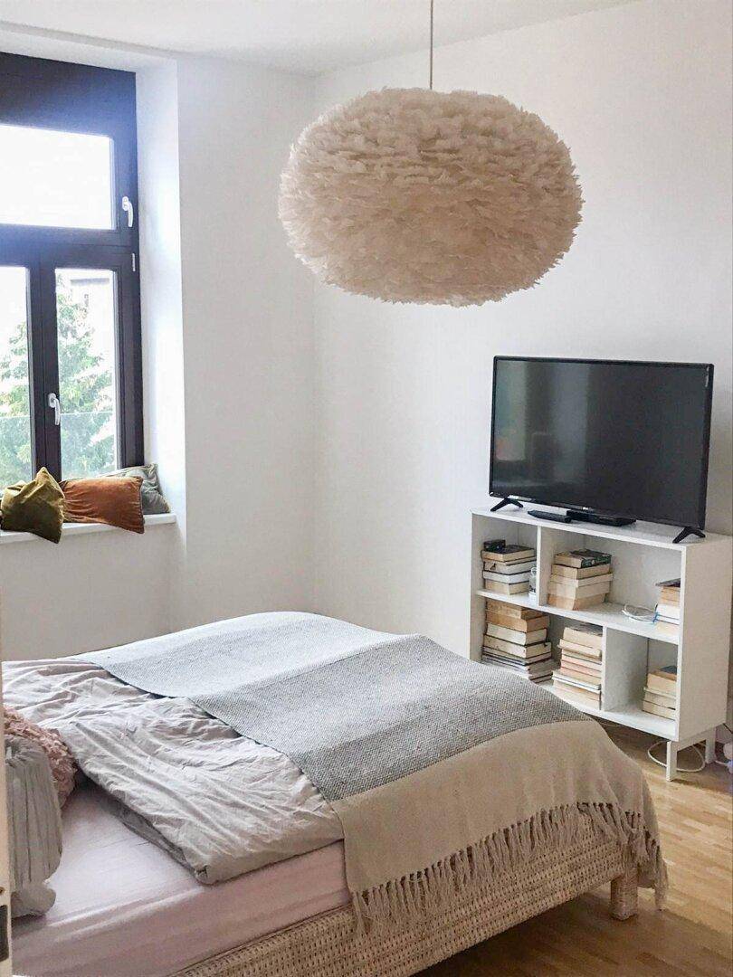 Gemütliches Schlafzimmer mit großem Fenster, hellem Federlampenschirm und Holzboden.