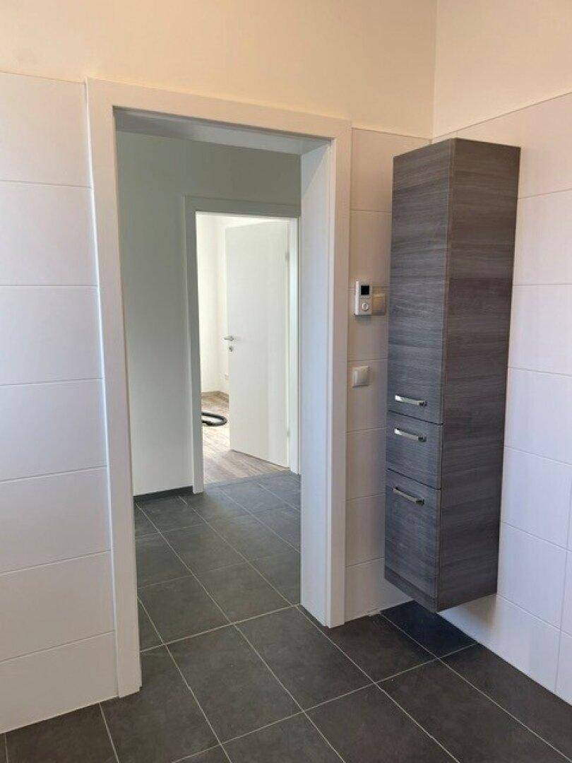 Heller Flur mit dunklen Fliesen und einem schmalen Hängeschrank, der zu weiteren Räumen führt.