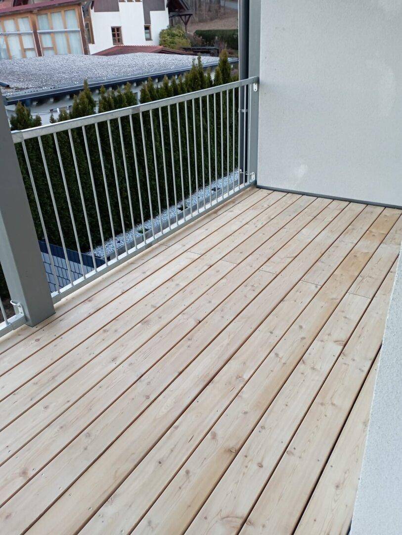 Geräumiger Balkon mit Holzdielenboden und Metallgeländer, umgeben von grünen Hecken.