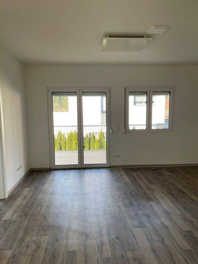 Heller Wohnraum mit Laminatboden und großen Schiebetüren, die zu einem Balkon mit grüner Bepflanzung führen.