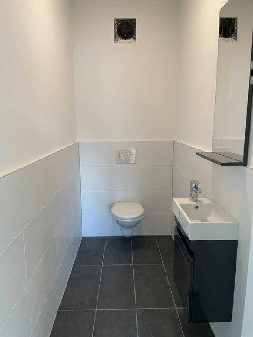 Modernes Gäste-WC mit dunklen Bodenfliesen, wandhängendem WC, kleinem Waschbecken und Abluftventilator.