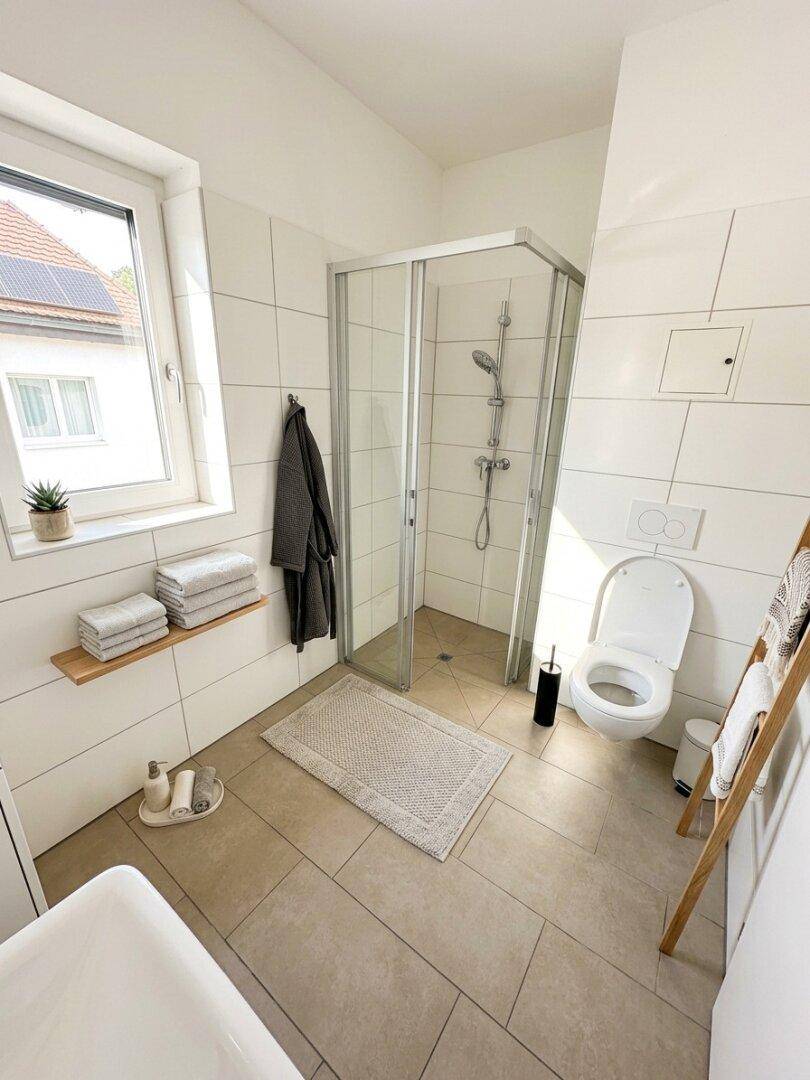 Modernes Badezimmer mit Dusche, WC, Fenster und hellen Fliesen, gut beleuchtet.