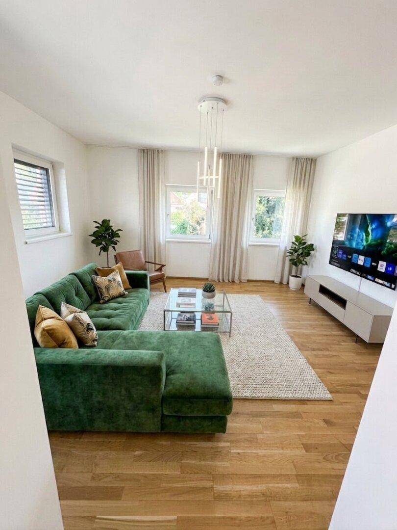 Geräumiges Wohnzimmer mit grünem Ecksofa, Holzboden und großen Fenstern, lichtdurchflutet.