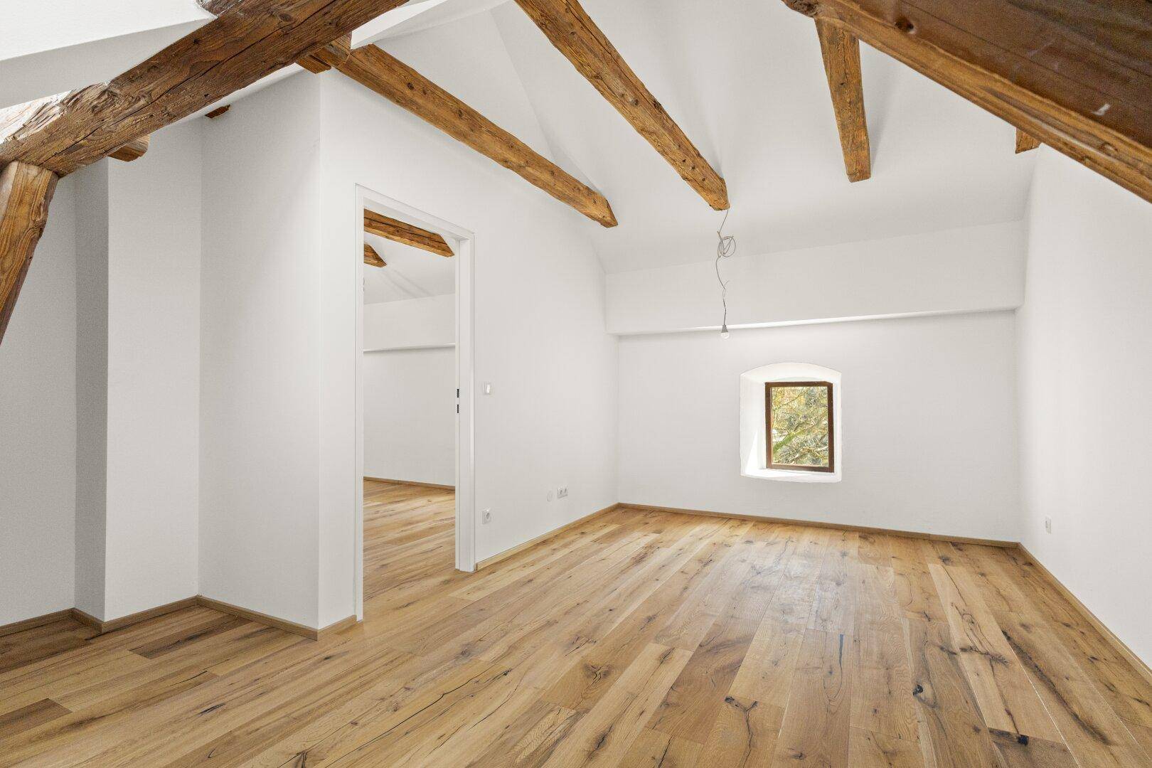 Heller, renovierter Dachraum mit sichtbaren Holzbalken, schrägen Decken und Dielenboden, bietet einen grünen Ausblick.
