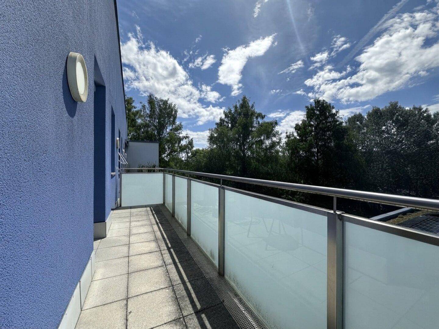 Heller Balkon mit Fliesenboden, Glasgeländer und Blick auf grüne Bäume und Himmel.
