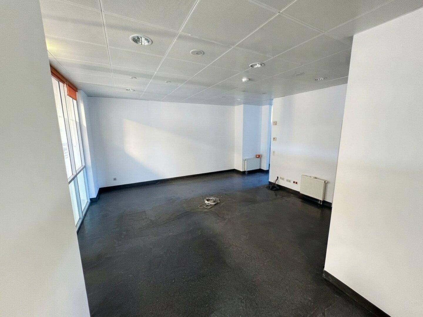 Leerer Büroraum mit weißen Wänden, dunklem Boden und viel Tageslicht durch große Fenster.
