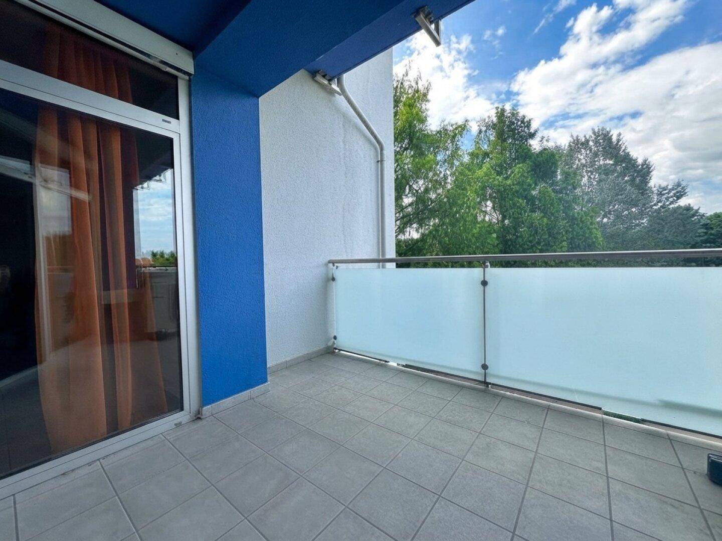 Geräumiger Balkon mit grauen Fliesen, Glasgeländer und blauer Fassade, umgeben von viel Grün.