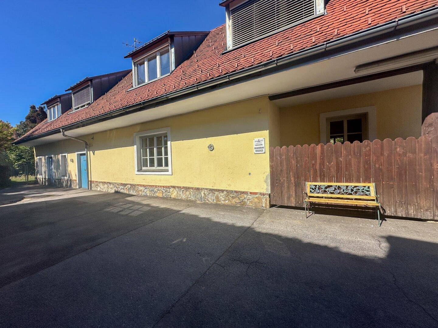 Eine gelbe Gebäudefassade mit rotem Ziegeldach, einem Holzzaun und einer Sitzbank im Freien.