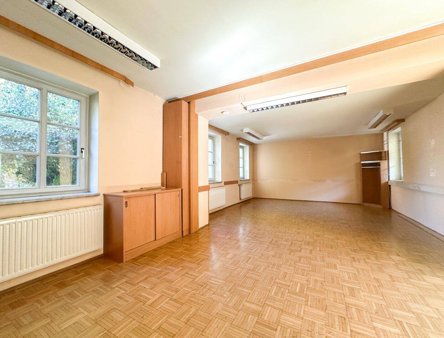 Geräumiger, leerer Büroraum mit Parkettboden, großen Fenstern und einem Holzschrank.
