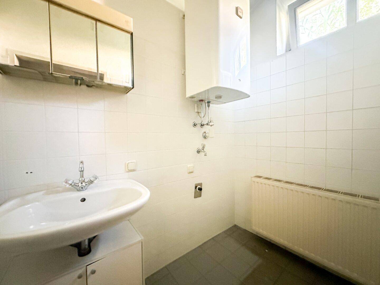 Ein kleines Badezimmer mit weißen Fliesen, einem Waschbecken, Warmwasserbereiter und einem Fenster.