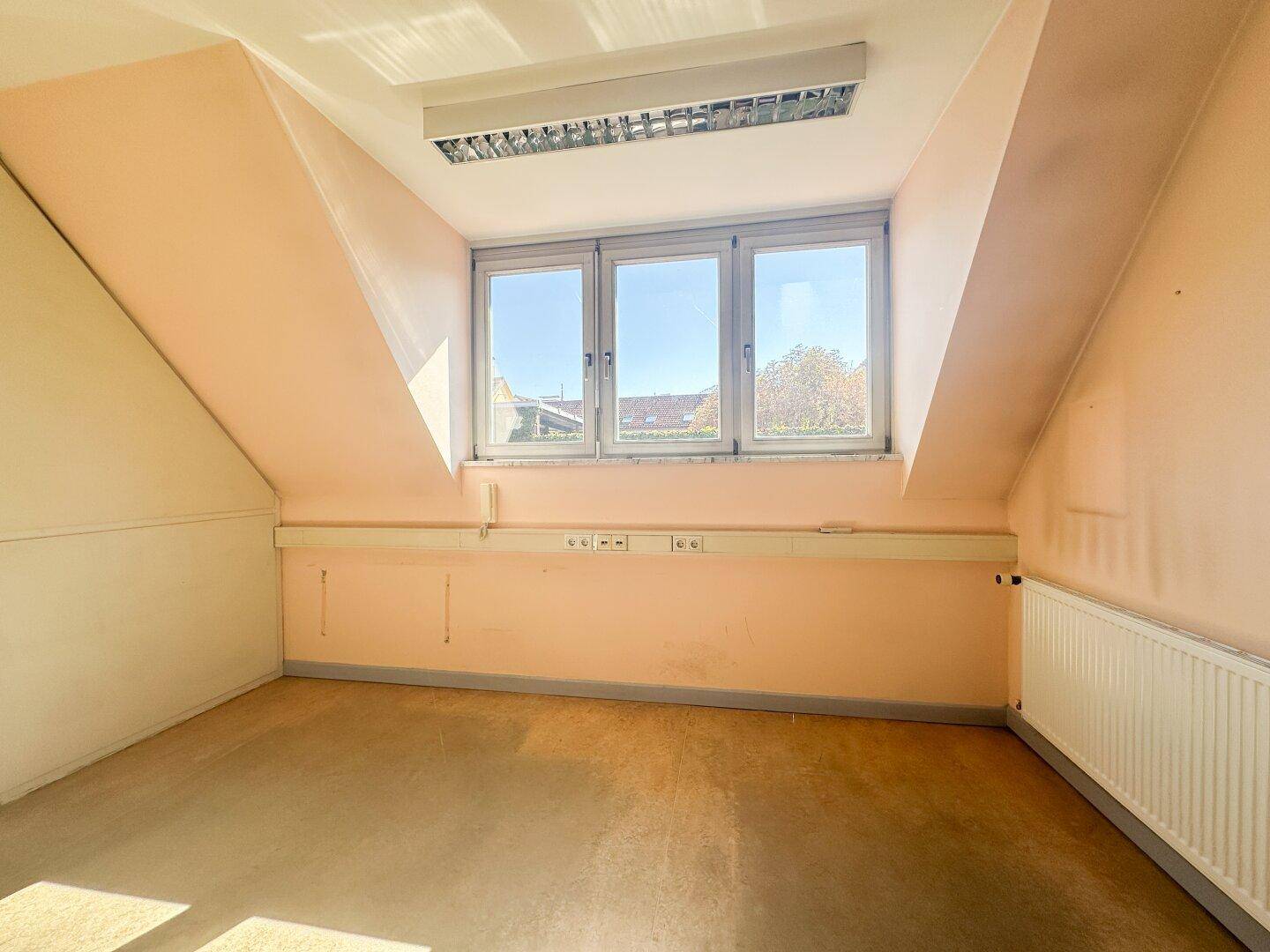 Ein heller, leerer Raum im Dachgeschoss mit Schrägen und einem breiten Fenster, ideal für ein Büro.