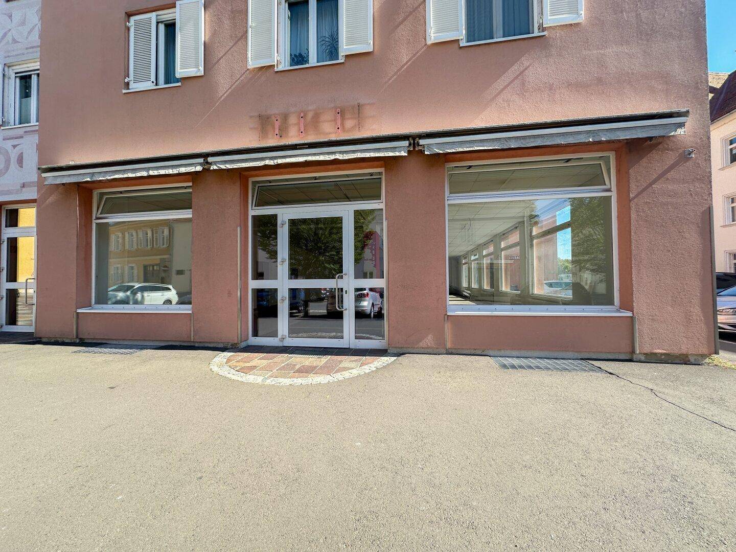 Vorderansicht eines Geschäftsgebäudes mit großen, leeren Schaufenstern und einem Eingangsbereich in der Sonne.