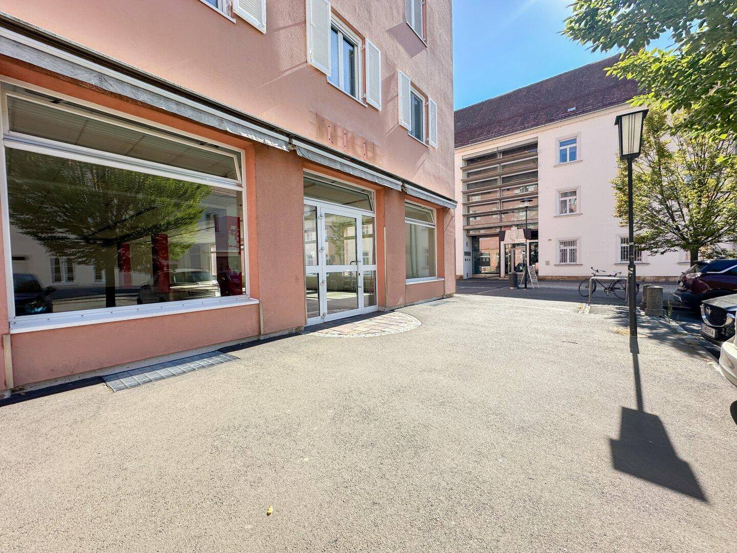 Seitenansicht eines Gebäudes mit großen Schaufenstern und einem gepflasterten Vorplatz bei Sonnenschein.