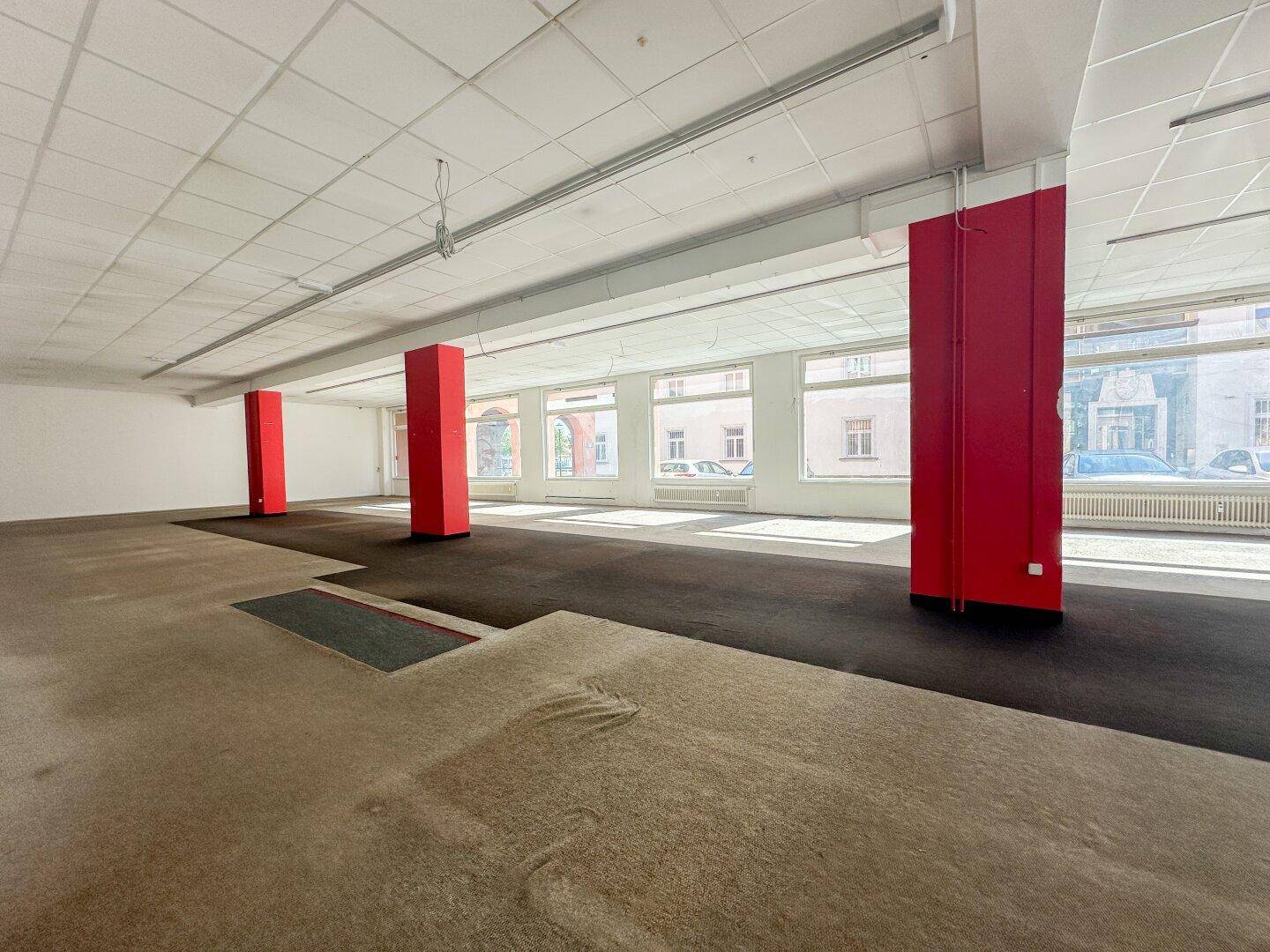 Geräumiger, leerer Gewerberaum mit roten Säulen, Teppichboden und großen Fenstern zur Straße hin.