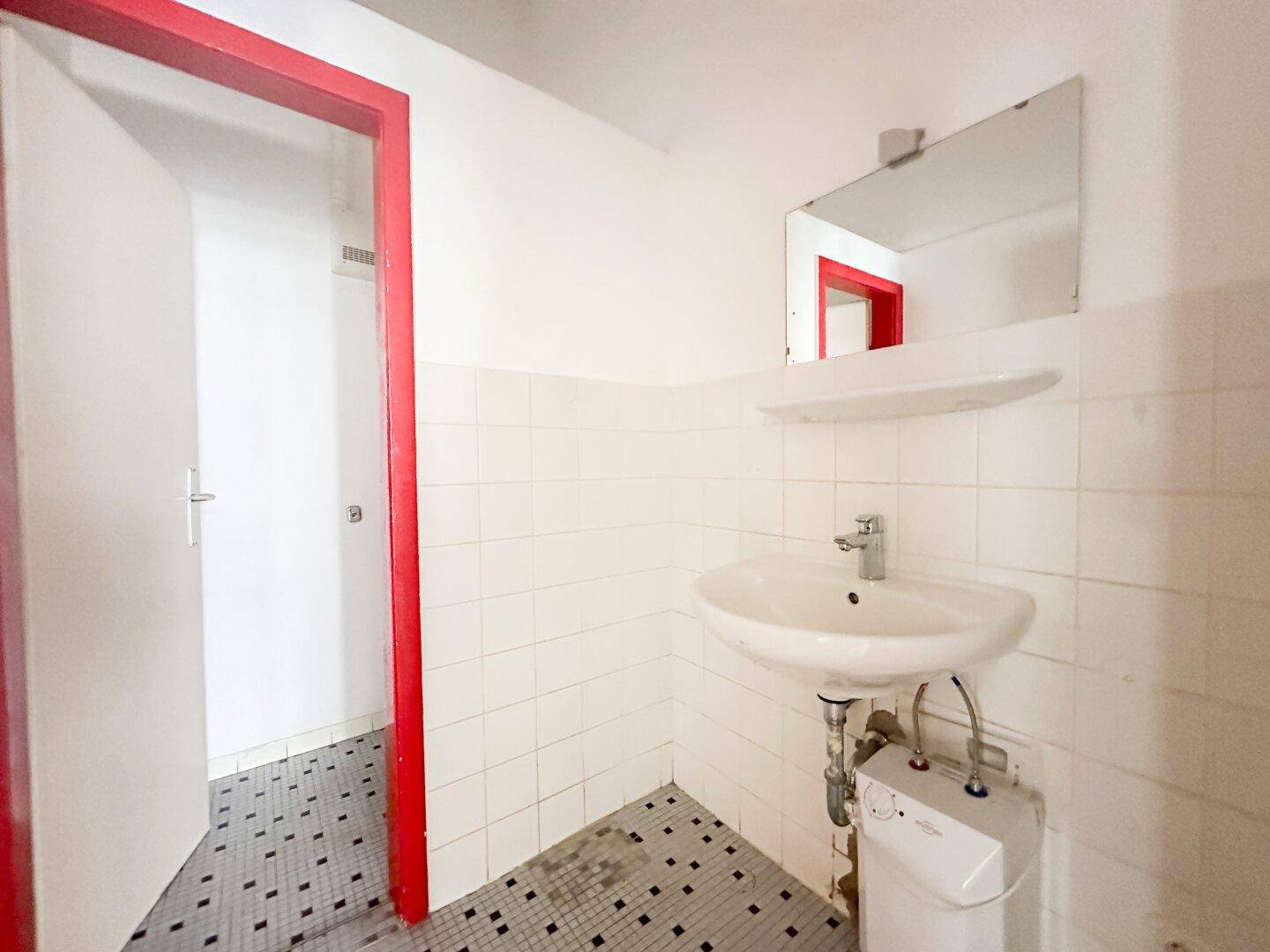 Einfache Toilette mit Waschbecken, Spiegel und weiß gefliesten Wänden, akzentuiert durch einen roten Türrahmen.