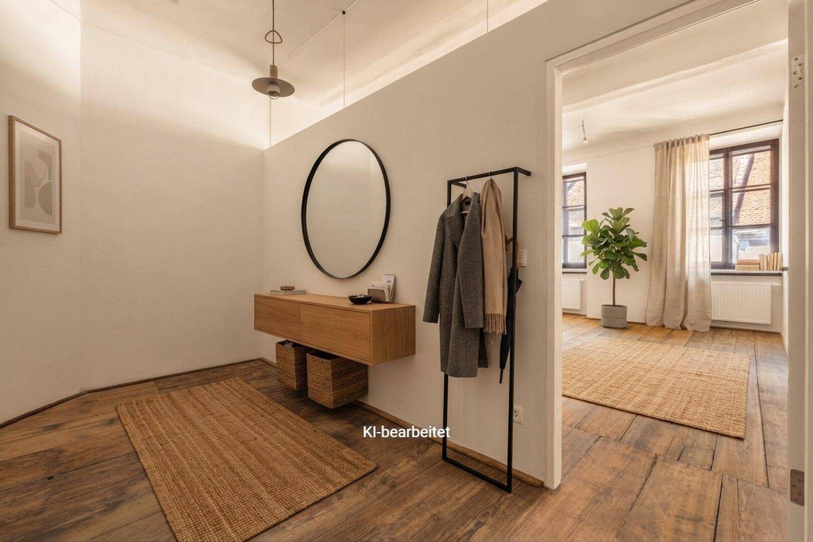 Heller Flur mit Holzboden, runder Spiegel und Garderobe, der in einen weiteren Raum führt.