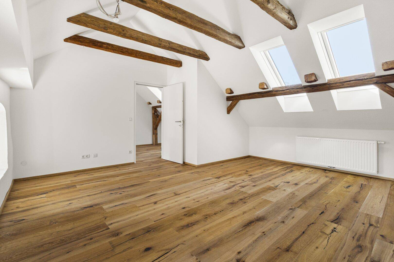 Heller, leerer Raum mit Holzbalken, Dachfenstern und Holzboden.