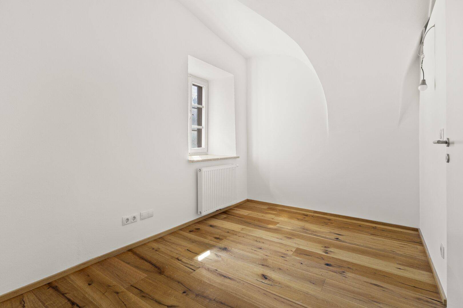 Kleiner, heller Raum mit Holzboden, Fenster und Heizkörper.