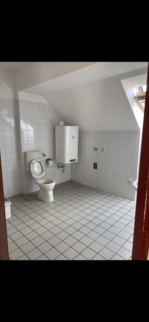 Badezimmer mit weißen Fliesen, Wand-WC und einem Warmwasserbereiter unter der Dachschräge.