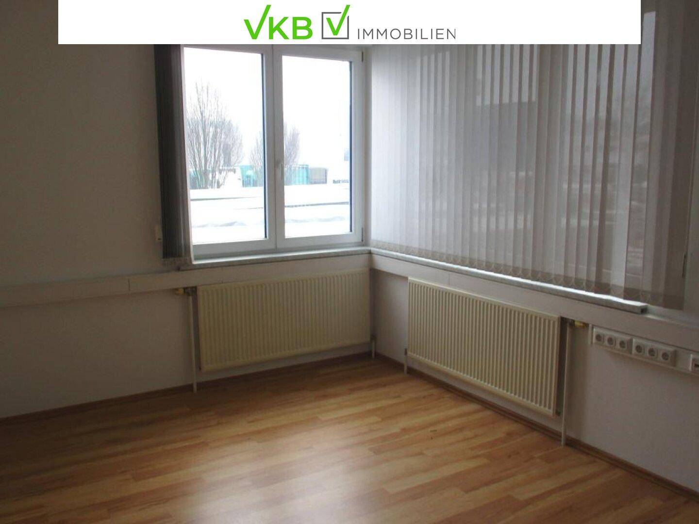 Leeres Büro mit Laminatboden und großem Fenster, das viel Tageslicht hereinlässt.