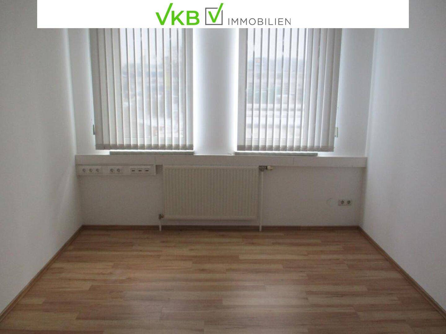 Helles, leeres Büro mit Laminatboden und zwei Fenstern, die für gute Belichtung sorgen.