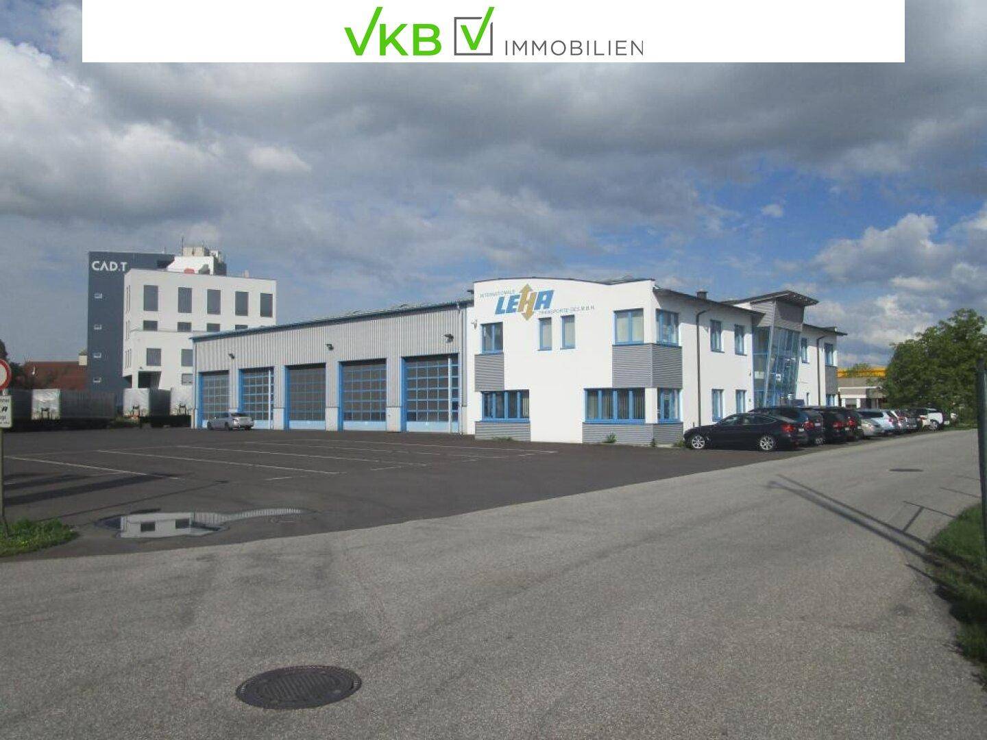 Gesamtansicht des Gewerbegebäudes mit Bürotrakt und Lagerhallen sowie großem asphaltierten Parkplatz.