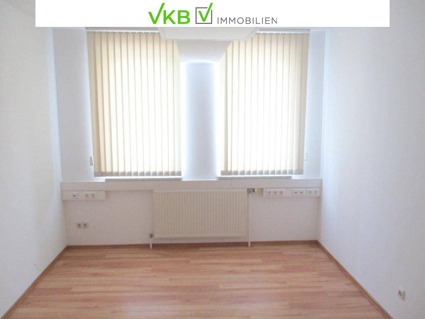 Helles Büro mit zwei Fenstern, vertikalen Jalousien, Laminatboden und Heizkörpern.