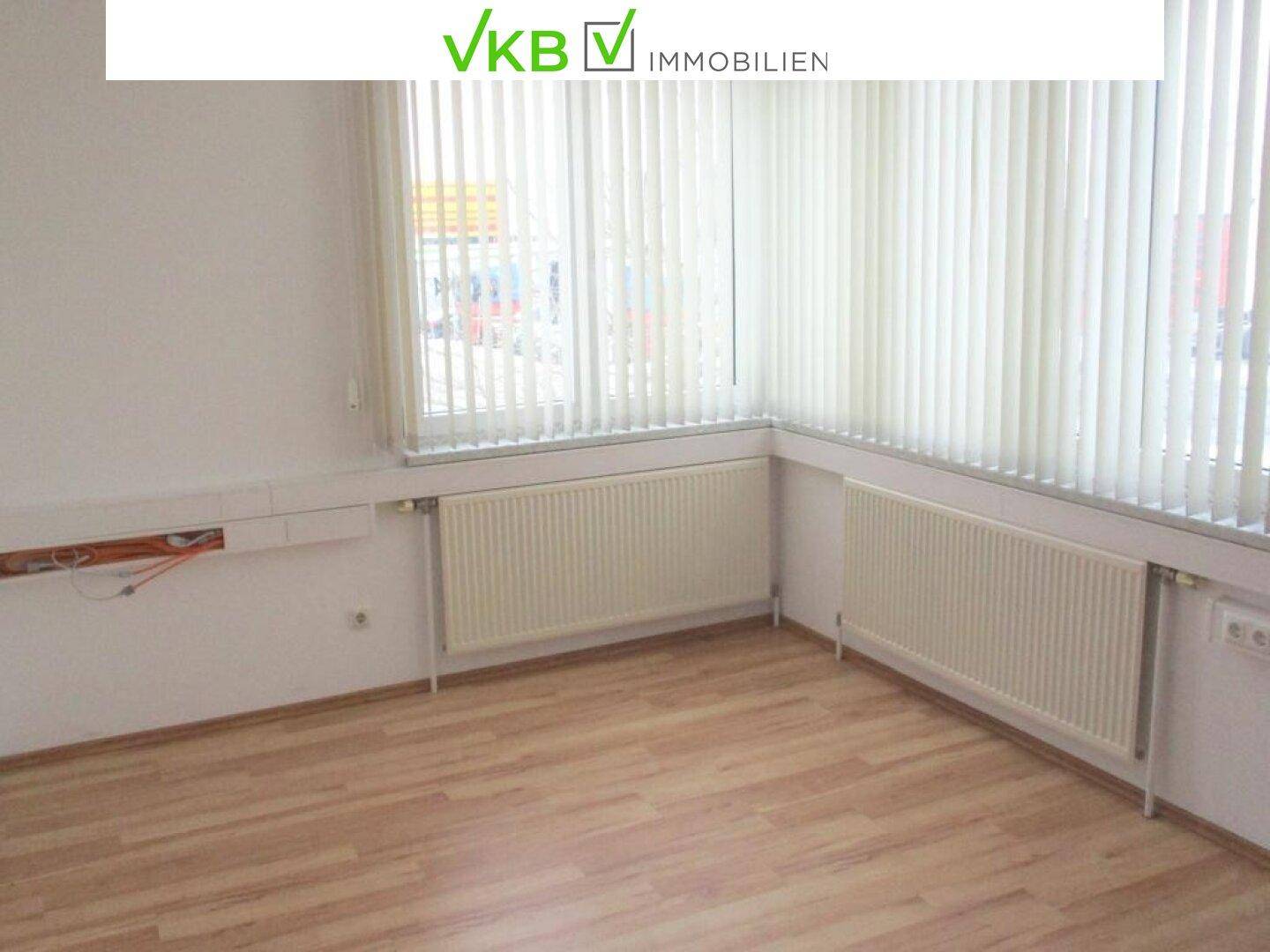 Helles Eckbüro mit Laminatboden, vertikalen Jalousien an den Fenstern und Heizkörpern.