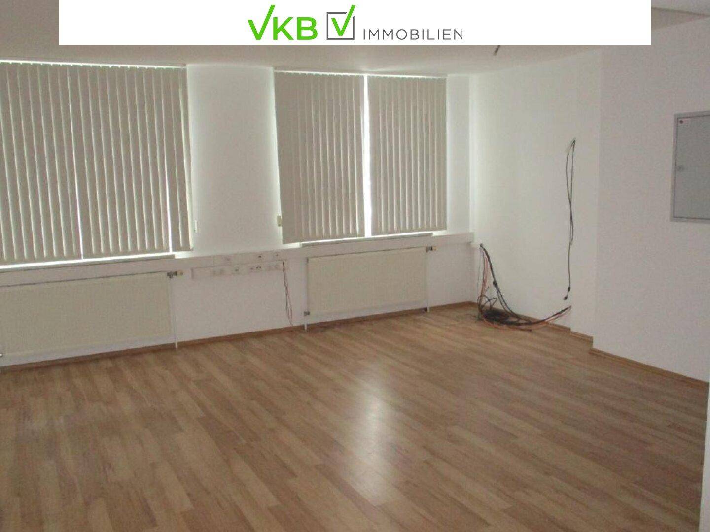 Geräumiges Büro mit hellem Laminatboden, großen Fenstern und Heizkörpern unter den Fensterbänken.