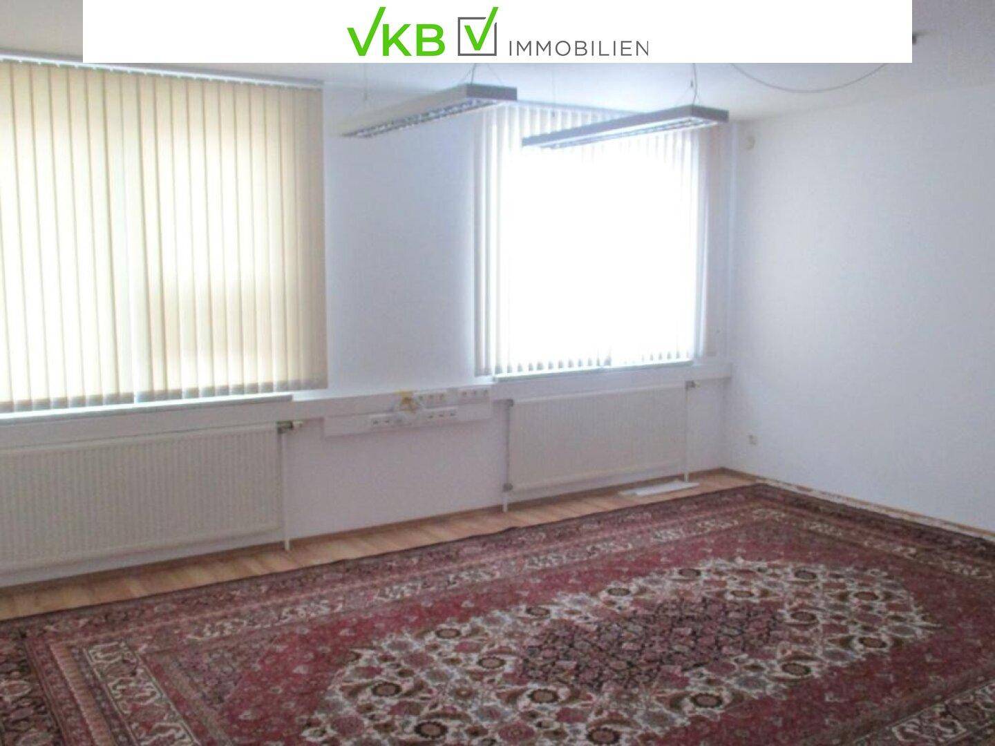 Büroraum mit Laminatboden, einem großen roten Teppich und Fenstern mit vertikalen Jalousien.