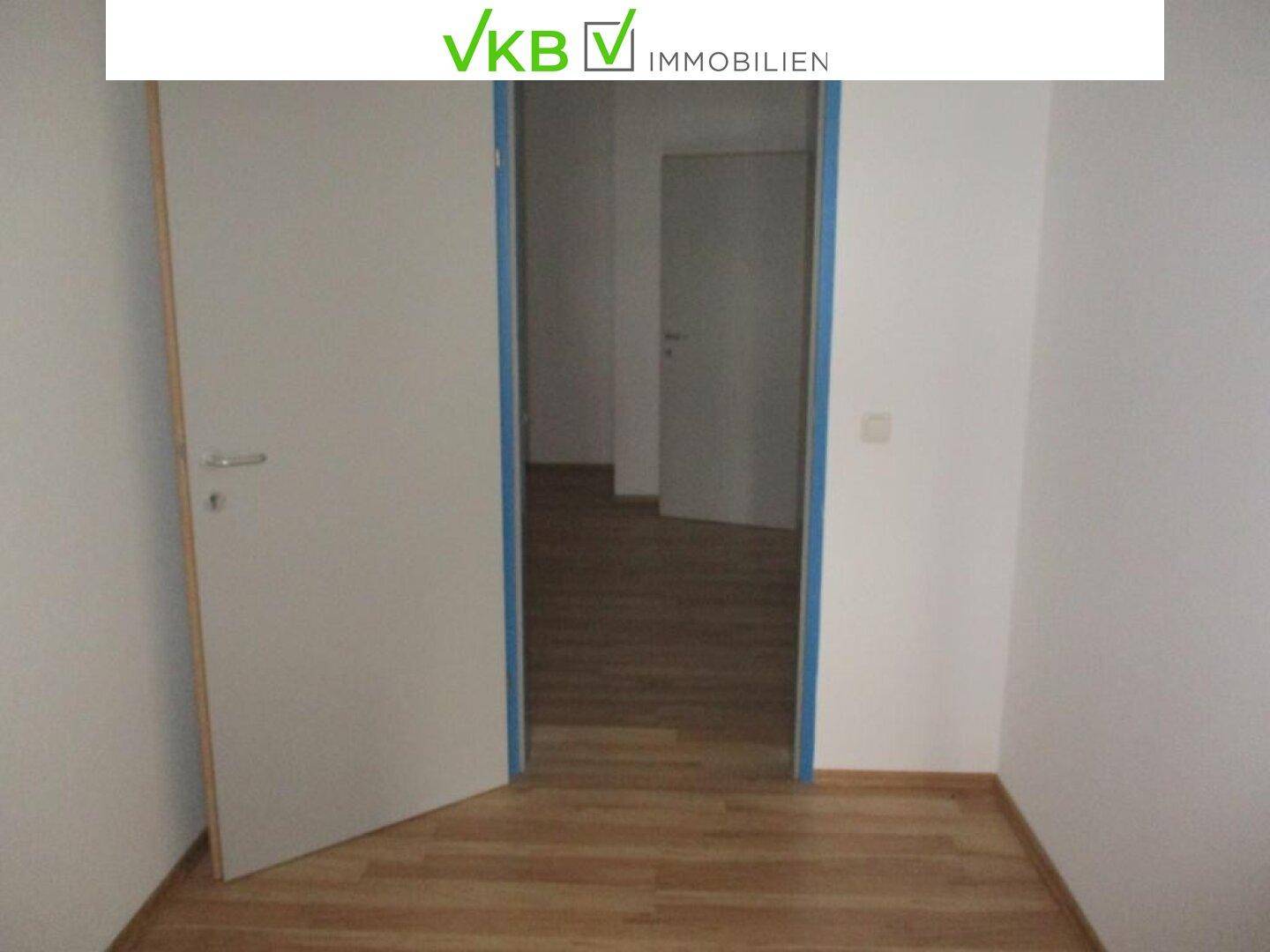Leerer Raum mit hellem Laminatboden und weiß gestrichenen Wänden, eine offene Tür führt in den Flur.
