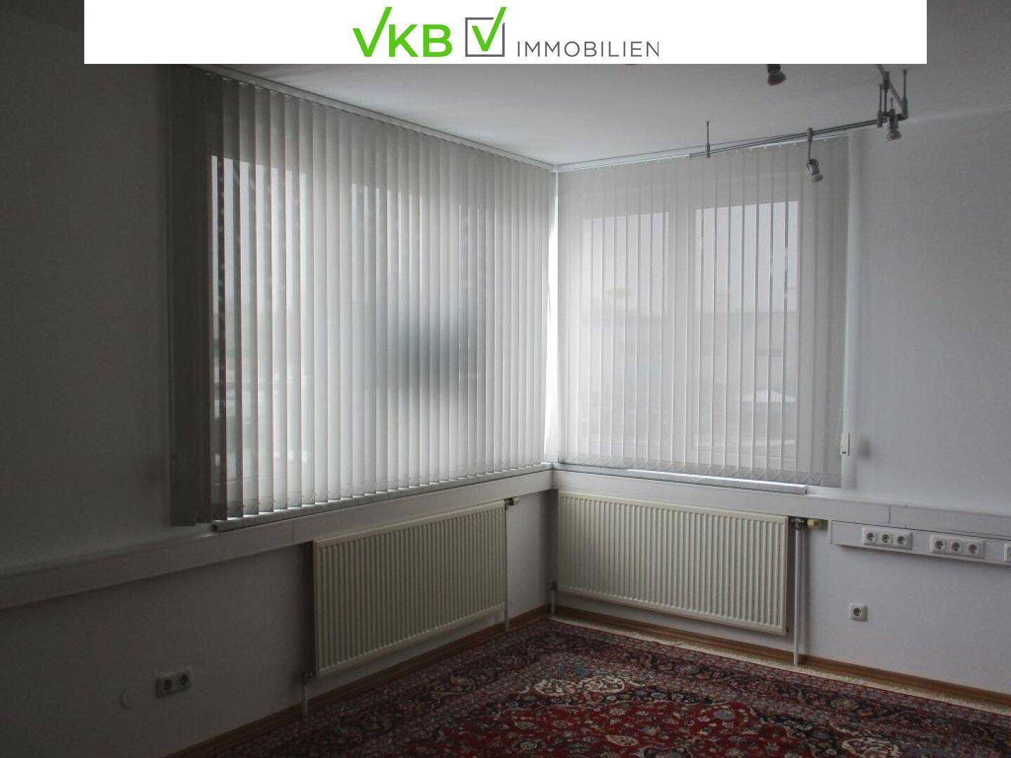 Raum mit vertikalen Jalousien vor den Fenstern, zwei Heizkörpern und einem gemusterten Teppich auf dem Boden.