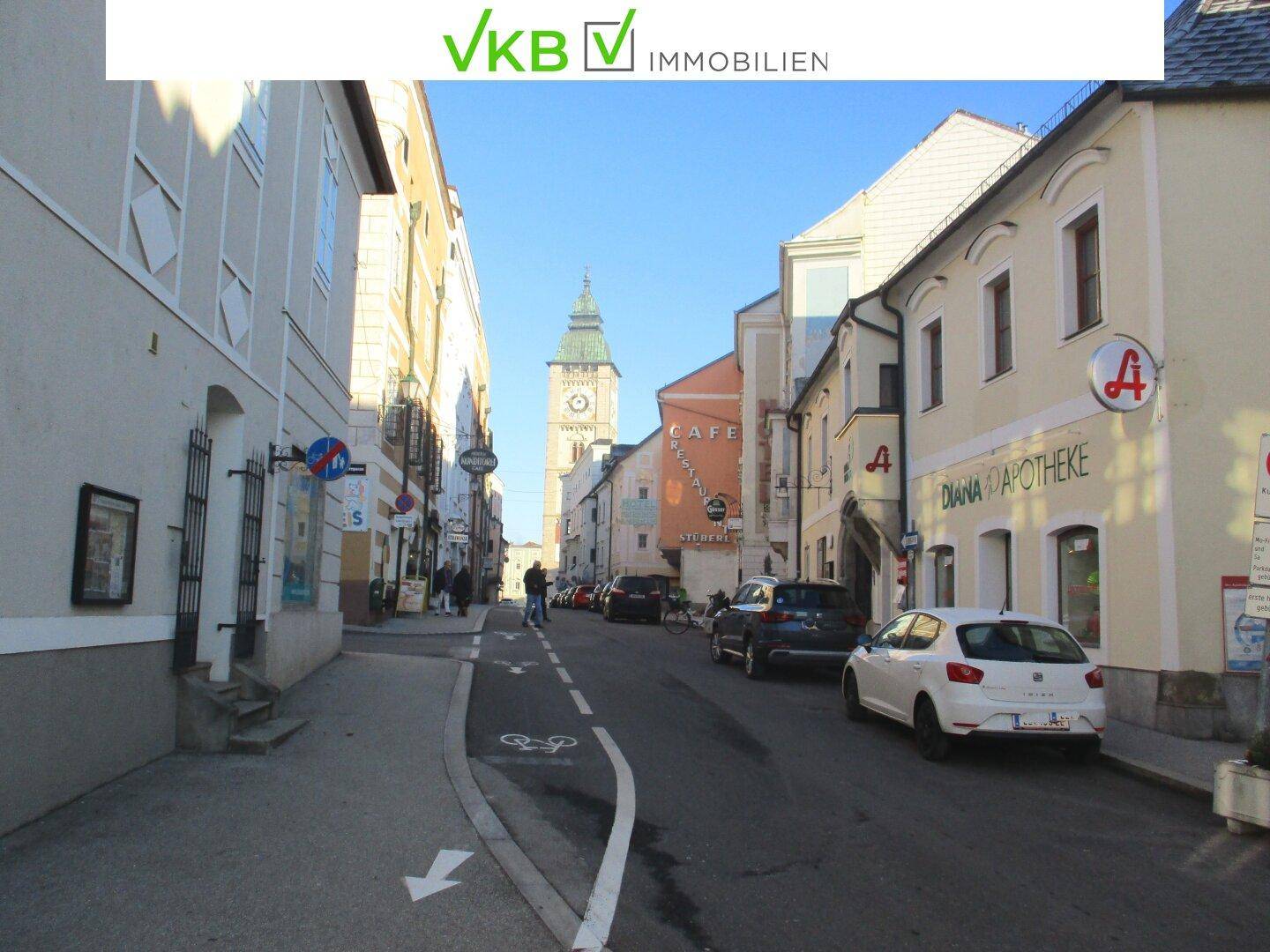 Blick auf eine belebte Straße in einer historischen Altstadt mit charmanten Gebäuden und einem markanten Uhrturm im Hintergrund unter blauem Himmel.
