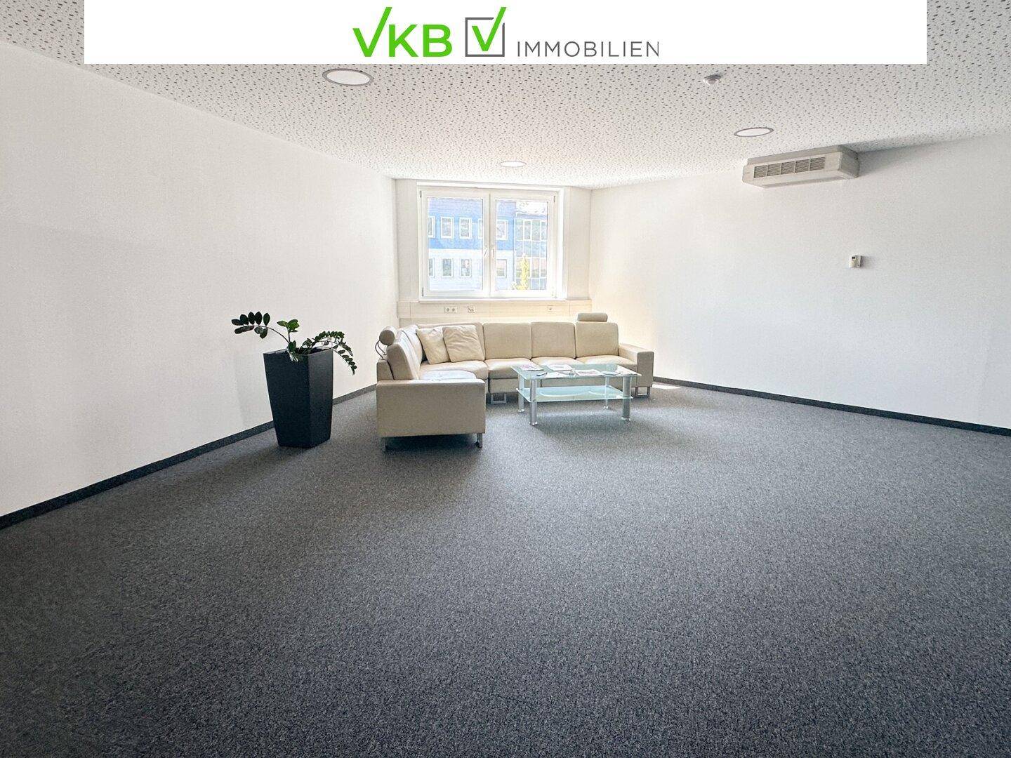 Großer, heller Büroraum mit beigem Sofa und grauem Teppichboden, ideal für eine Lounge oder Wartebereich.