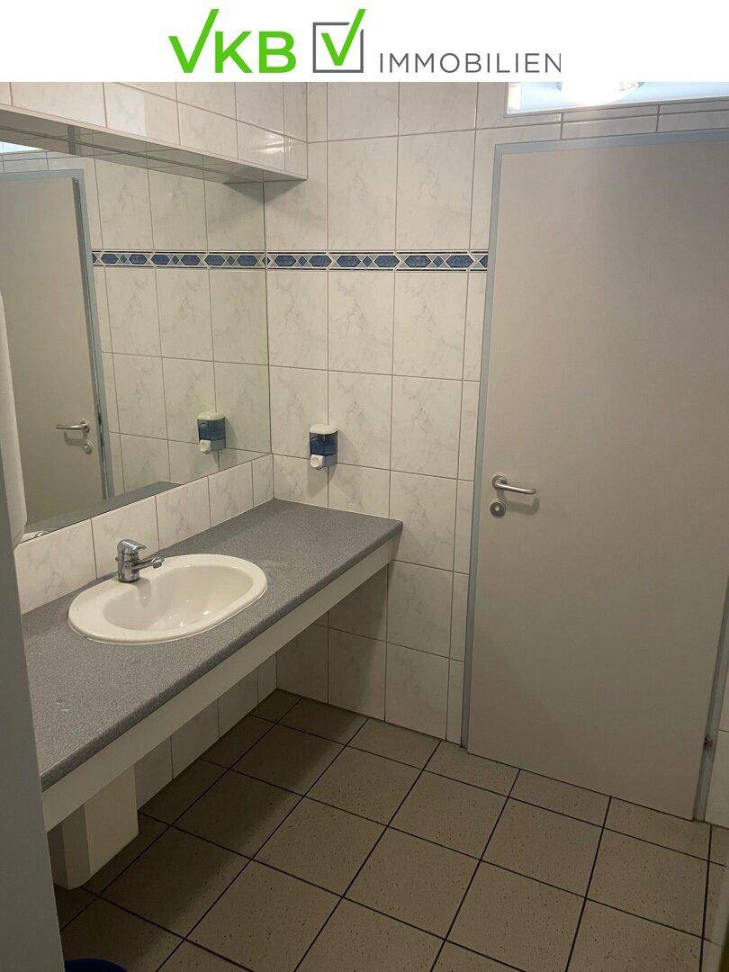Öffentliche Toilette mit Waschbecken, großem Spiegel und weißen Fliesen mit blauer Bordüre.