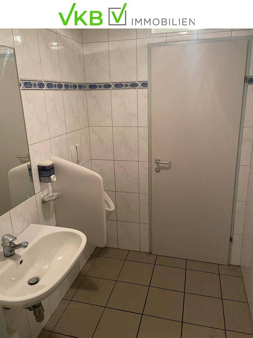 Herren-Toilette mit Waschbecken, Urinal und weißen Fliesen mit blauer Bordüre.
