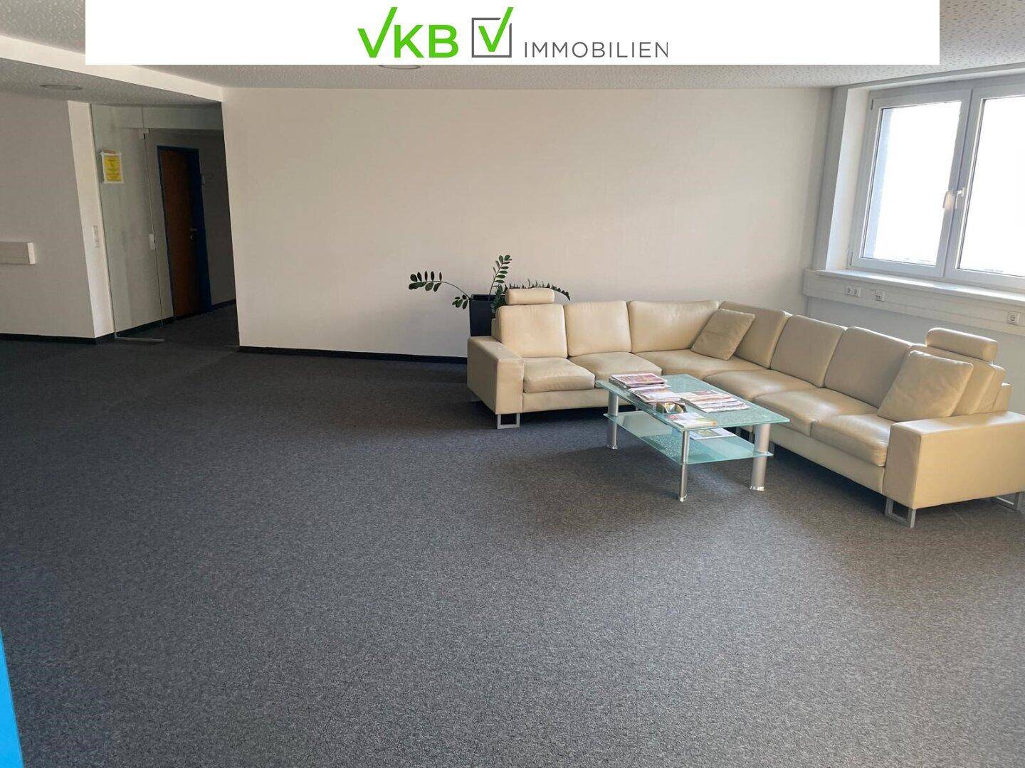 Geräumiger Bürobereich mit beigem Ecksofa, Glastisch und großen Fenstern, die viel Tageslicht hereinlassen.