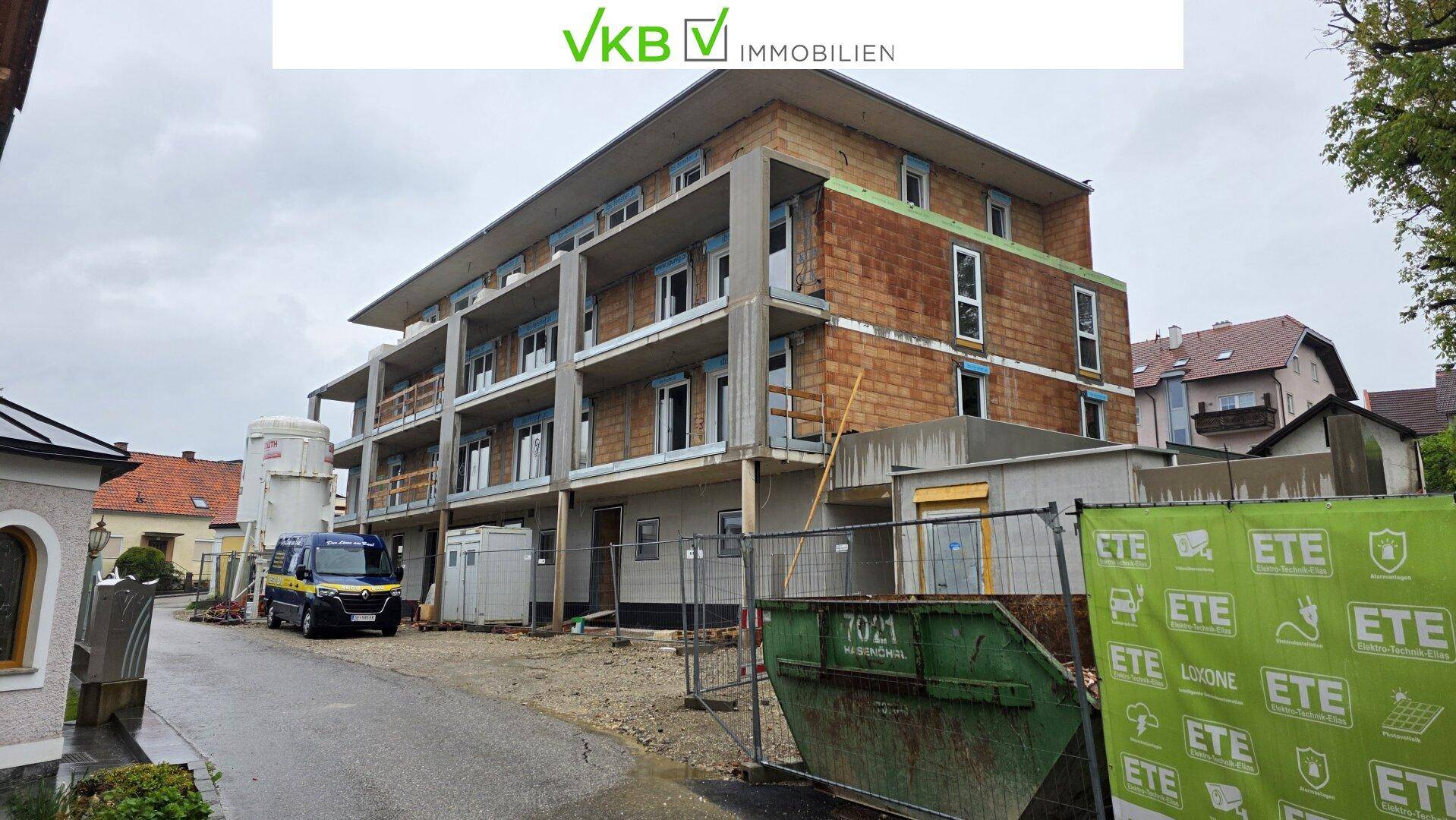 Baustelle des Neubaus mit teilweise verputzter Fassade, Baucontainern und einem Bauschuttcontainer.