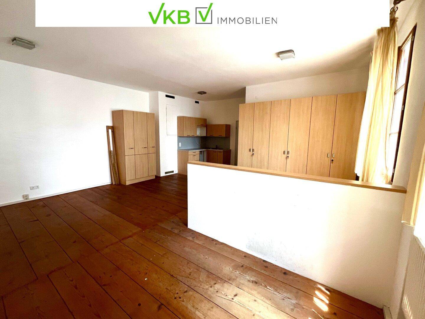 Offener Wohnbereich mit Holzdielenboden und einer integrierten Küchenzeile im Hintergrund.