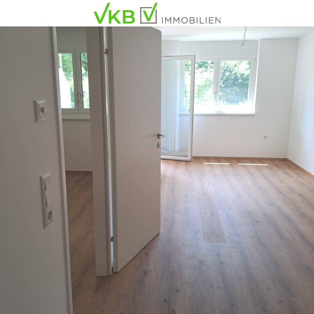 Heller Wohnraum mit Laminatboden und großen Fenstern, die viel Tageslicht hereinlassen.