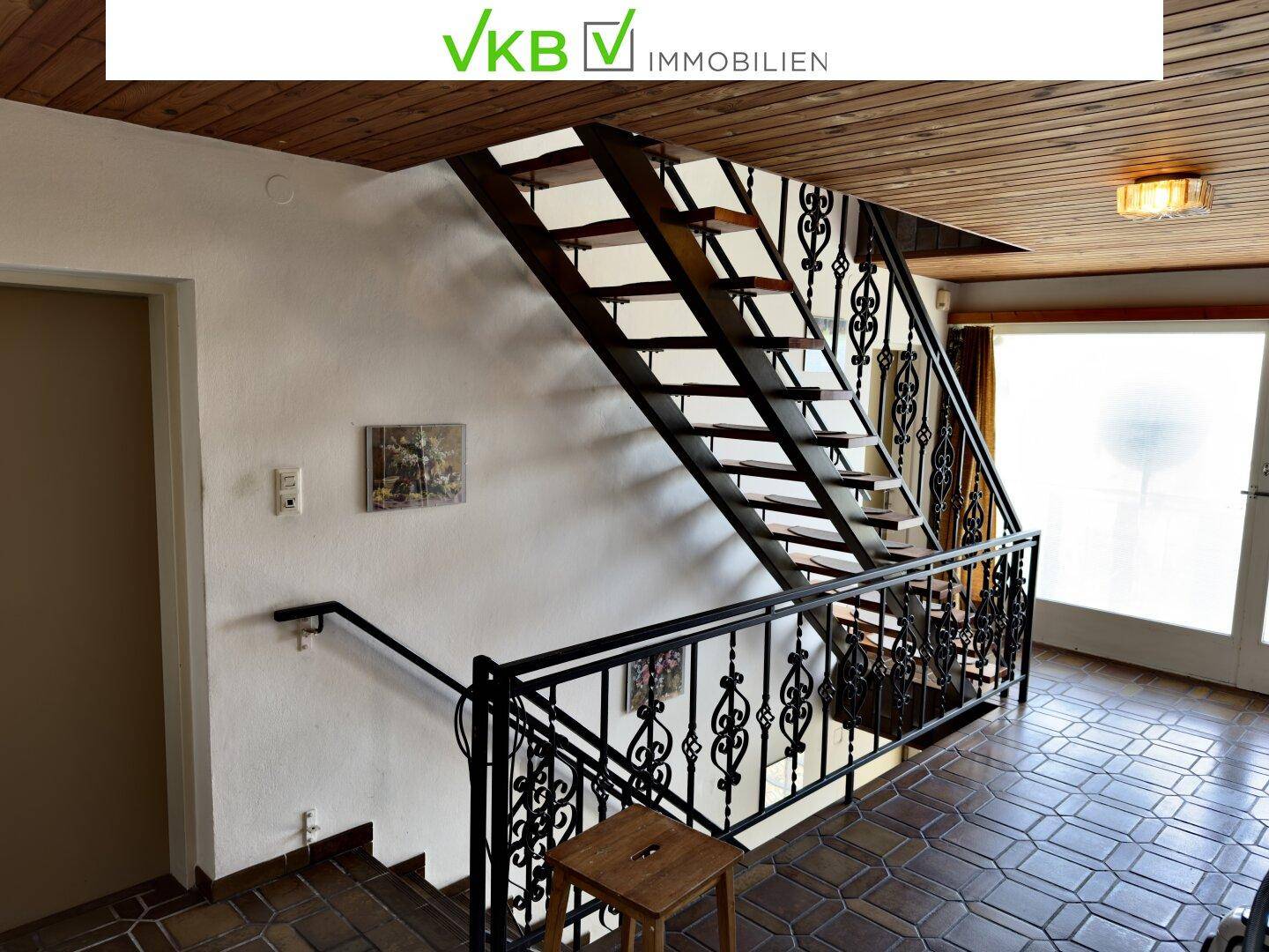 Treppenhaus mit schmiedeeisernem Geländer und Holzstufen, ergänzt durch eine Holzdecke und Fliesenboden.