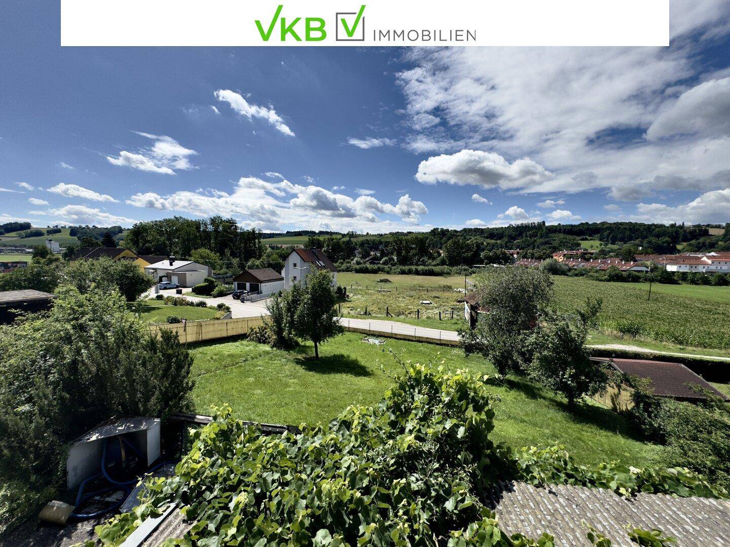 Weitläufiger Blick über grüne Felder und ländliche Häuser unter einem blauen Himmel mit Wolken.