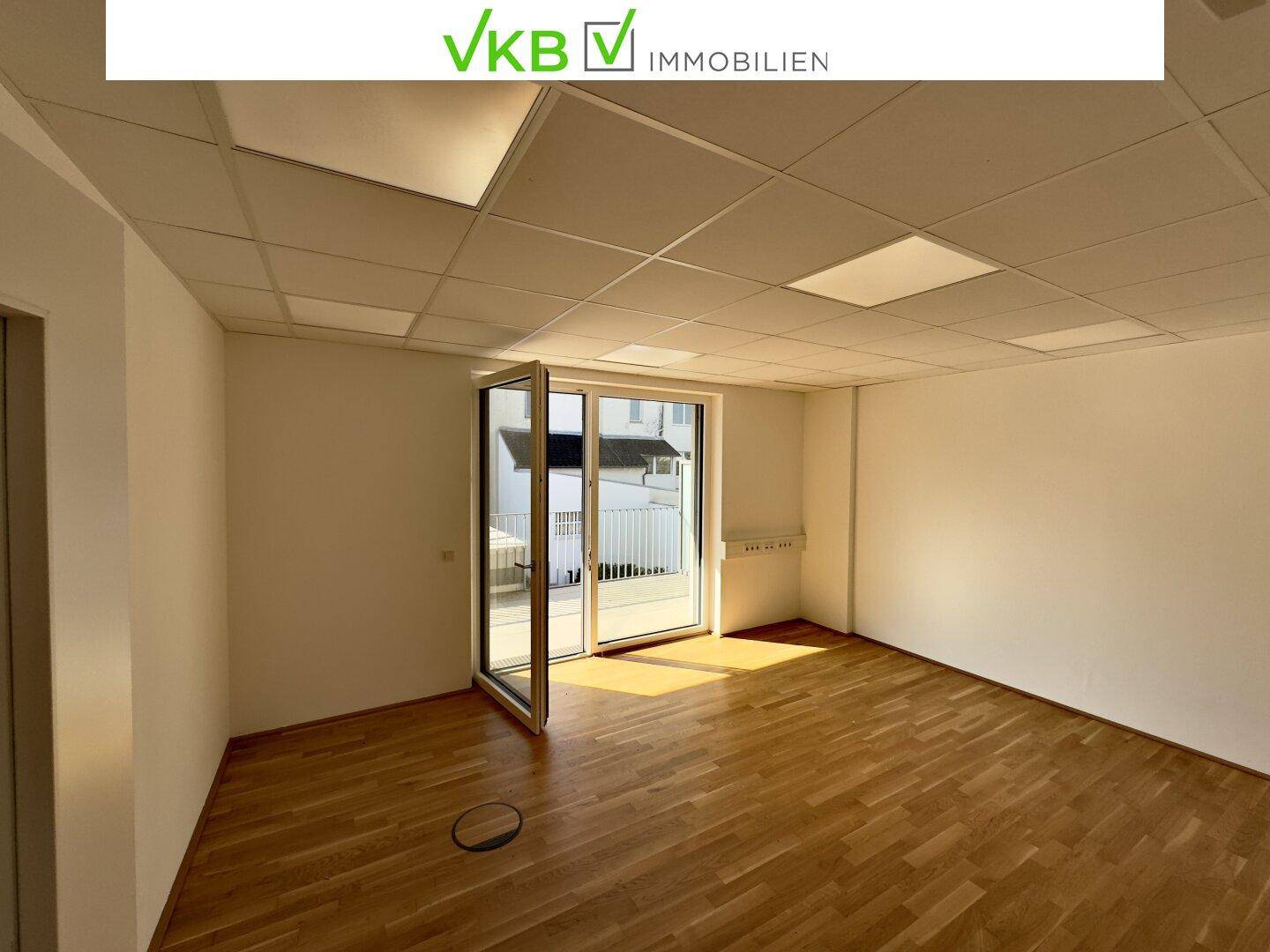 Heller Büroraum mit Parkettboden, weißen Wänden und Zugang zu einem Außenbereich.