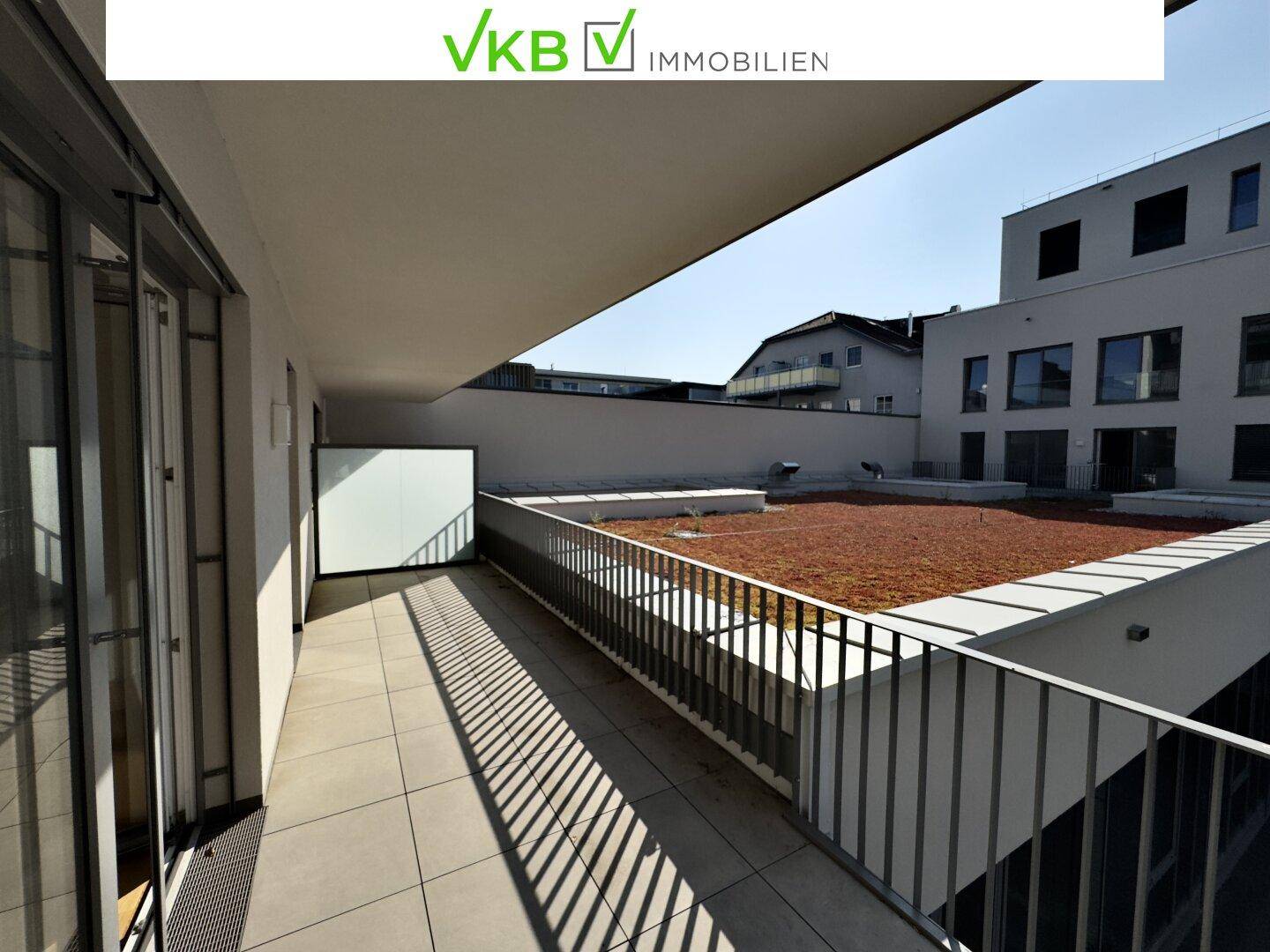 Geräumiger Balkon mit Fliesenboden und Blick auf ein begrüntes Dach und umliegende Gebäude.